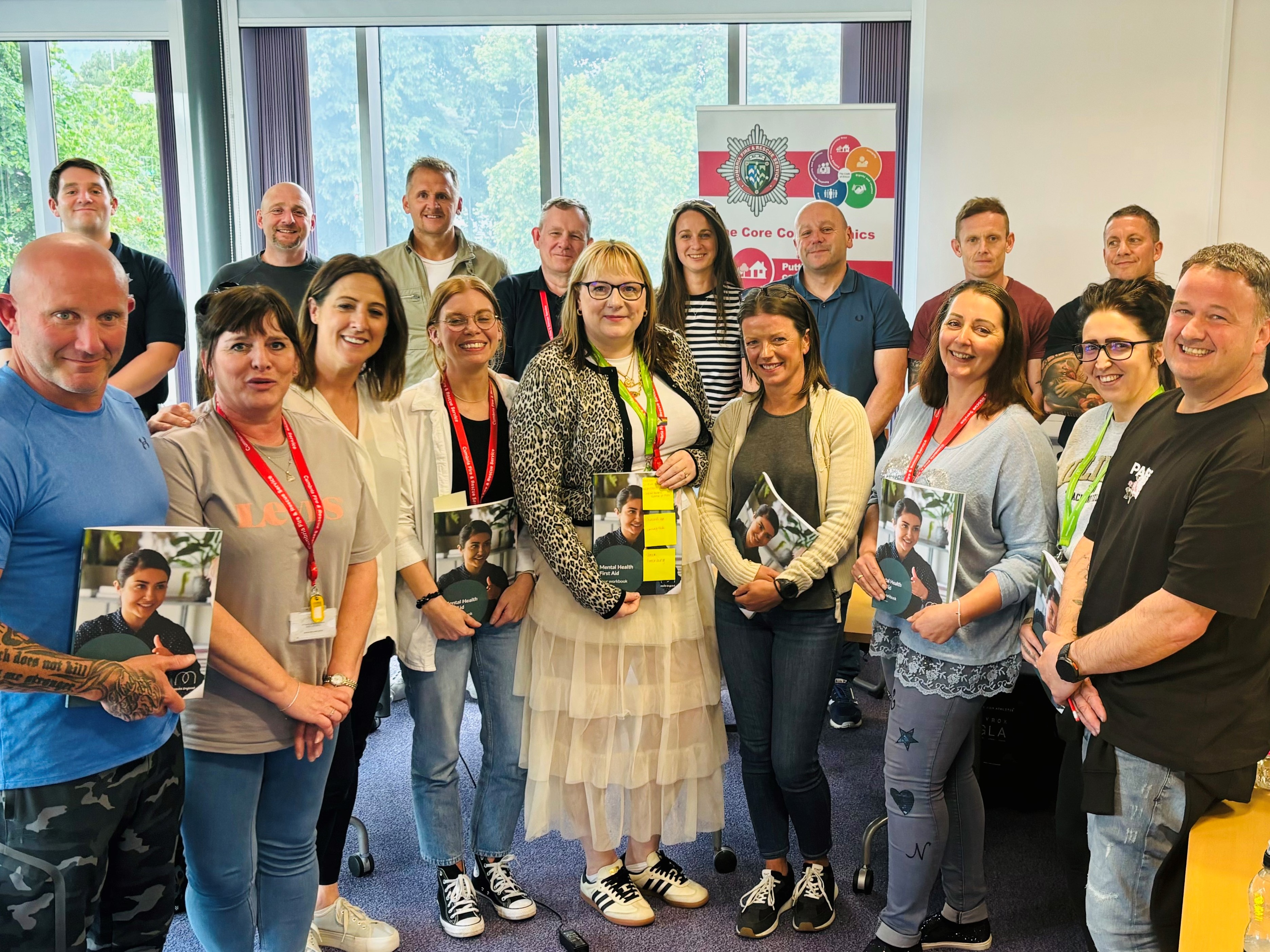 Image of adults standing in a group smiling at the camera with Mental Health First Aider workbooks in their hands