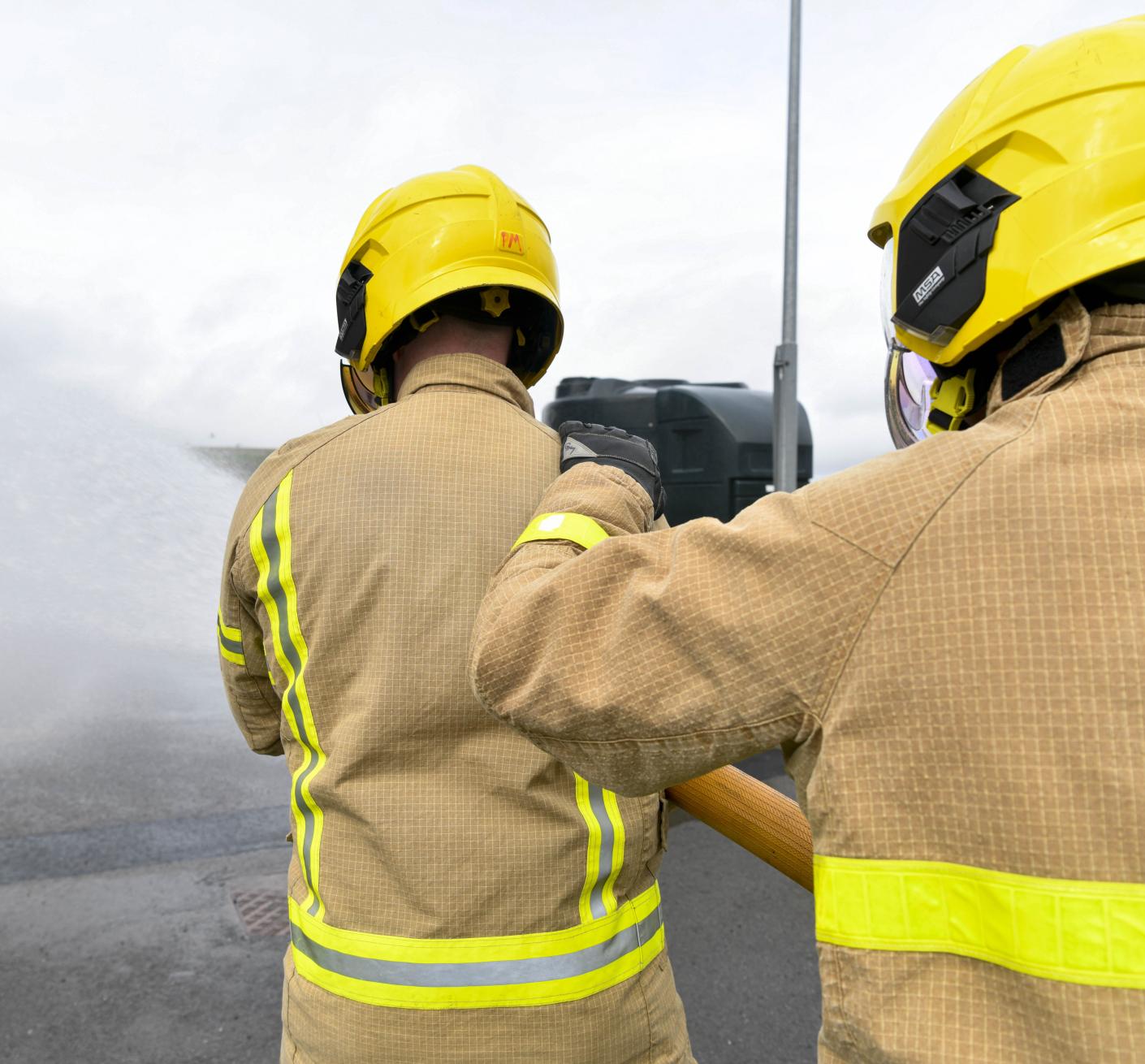 Two Firefighters in full PPE, the first one is holding a hose that is projecting water